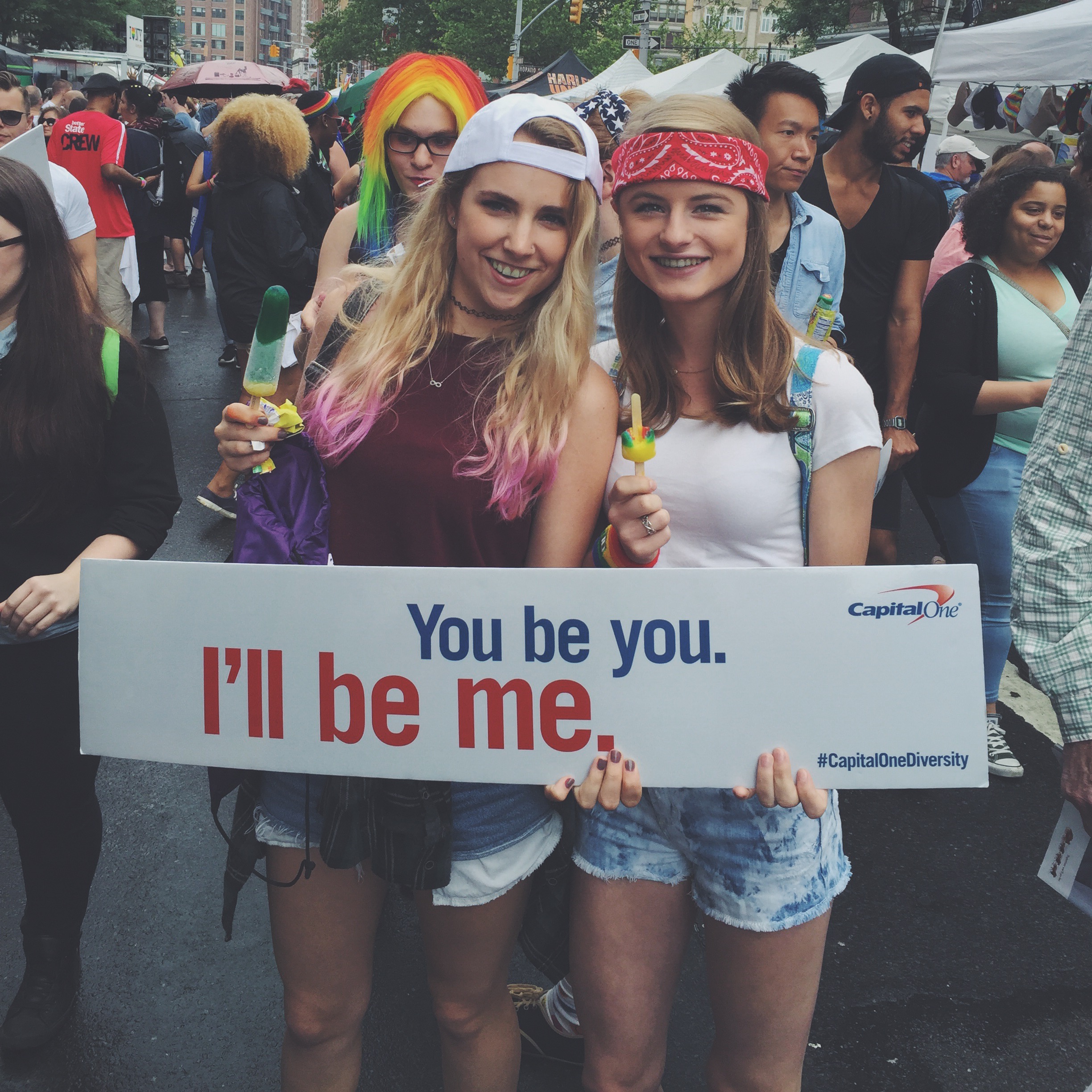 two friends holding a "you be you. I'll be me." sign created by Capital One.They are at the NYC Gay Pride Parade